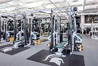 Michigan State Football Weight Room - Weight Rack View of the weight racks that are available in the Michigan State football weight room facility. Each weight rack has an individual REGUPOL crash zone that allows for increased durability and features an inlaid Spartan logo.