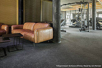 Seating area with brown sofas in a fitness studio, training equipment can be seen in the background. The flooring is dark grey.