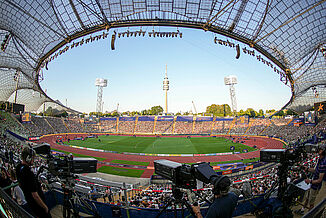 Munich Olympic Stadium during the 2022 European Athletics Championships – red track, TV crews, and international competition flair. Interior view of Munich Olympic Stadium during the 2022 European Athletics Championships. The red synthetic running track and central field are ready for competition, with broadcast cameras and spectators capturing the live atmosphere.