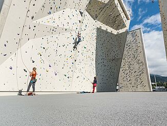 Outdoor climbing area with fall protection floor. The flooring is grey. A few people can be seen climbing on the climbing wall, with belay partners standing on the ground.