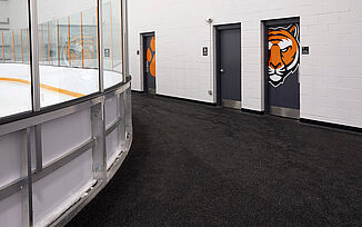 Curved Perspective of Thayer Sports Center Hockey Rink An angled view of the walkway around the Thayer Sports Center hockey rink, highlighting the curved shape of the rink and a doorway with the tiger logo leading into locker rooms. REGUPOL aktiv roll flooring was used throughout the walkway to ensure safety measures and durability are met for athletes.
