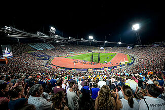 Crowded Munich Olympic Stadium during the 2022 European Athletics Championships. Lit red running track and central field at night. Munich Olympic Stadium during the 2022 European Athletics Championships at night. The red synthetic running track is illuminated, with thousands of spectators watching international track and field competitions under floodlights