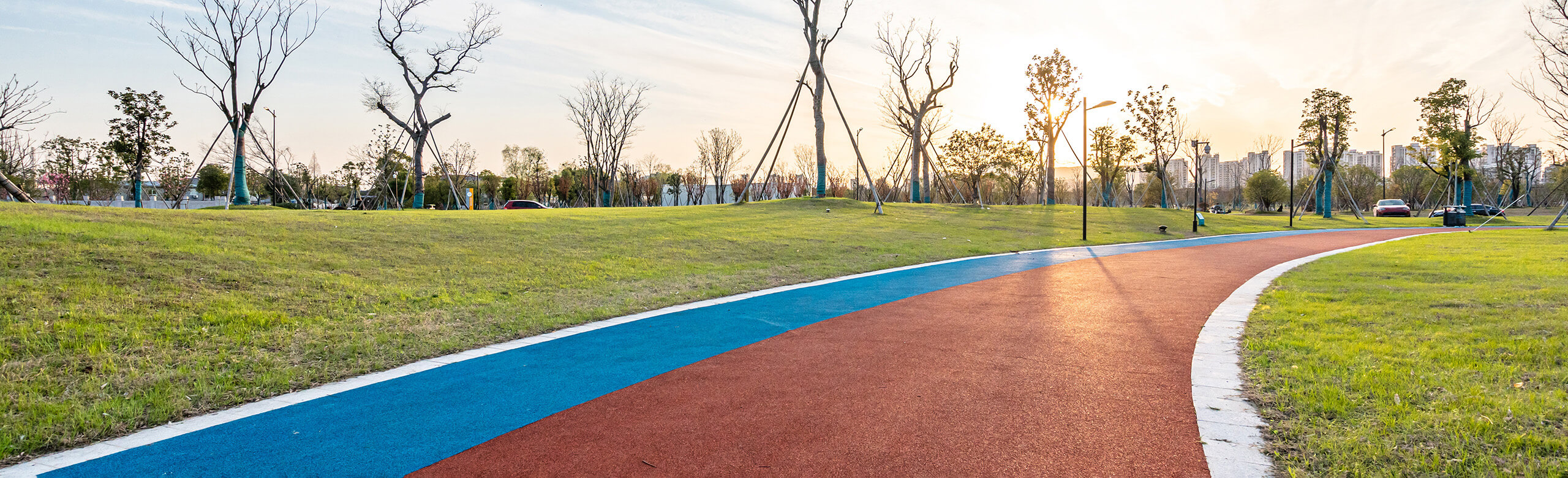 Outdoor synthetic jogging track with red and blue lanes, integrated into a green park landscape at sunset. The surface is made of elastic, water-permeable synthetic material – ideal for joint-friendly running and fitness training.0