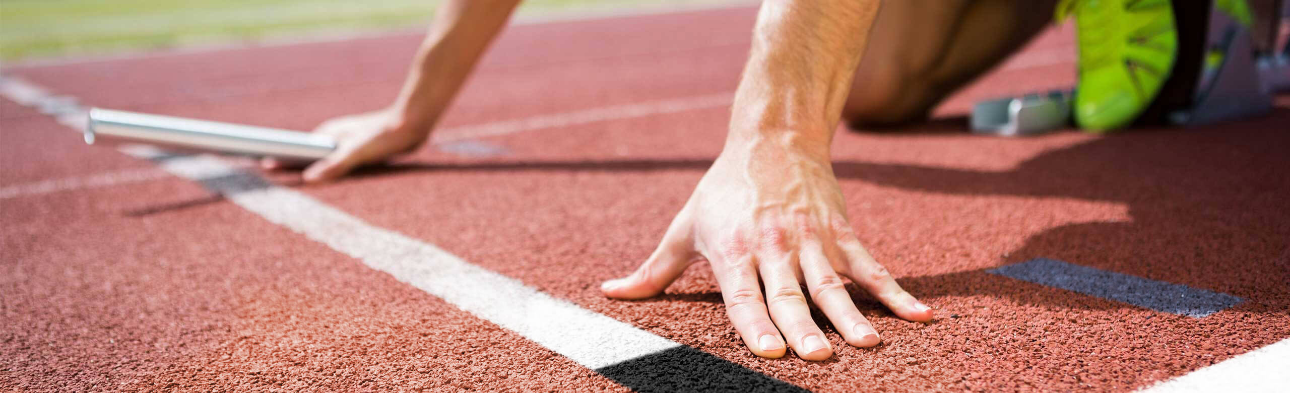 Close-up of an athlete in starting position on a red REGUPOL track, holding a relay baton, with starting blocks and spikes.