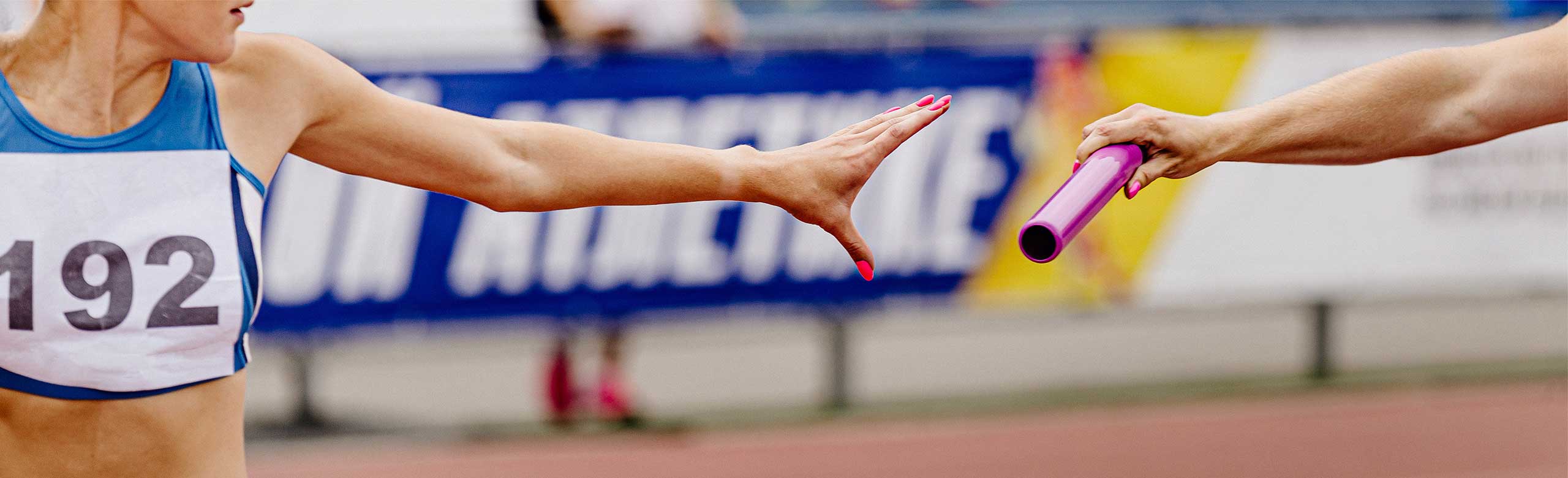 Close-up of a baton handover in a track and field relay race. A female runner in blue sportswear reaches out to receive the purple baton from her teammate. The background shows a running track and a blurred advertisement board.