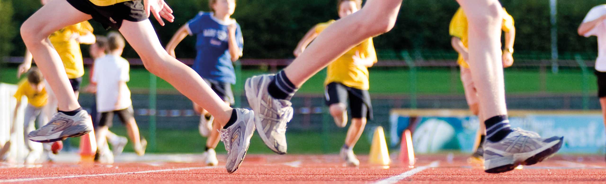 Children and young athletes running and playing on a red synthetic track under sunny skies at the stadium.