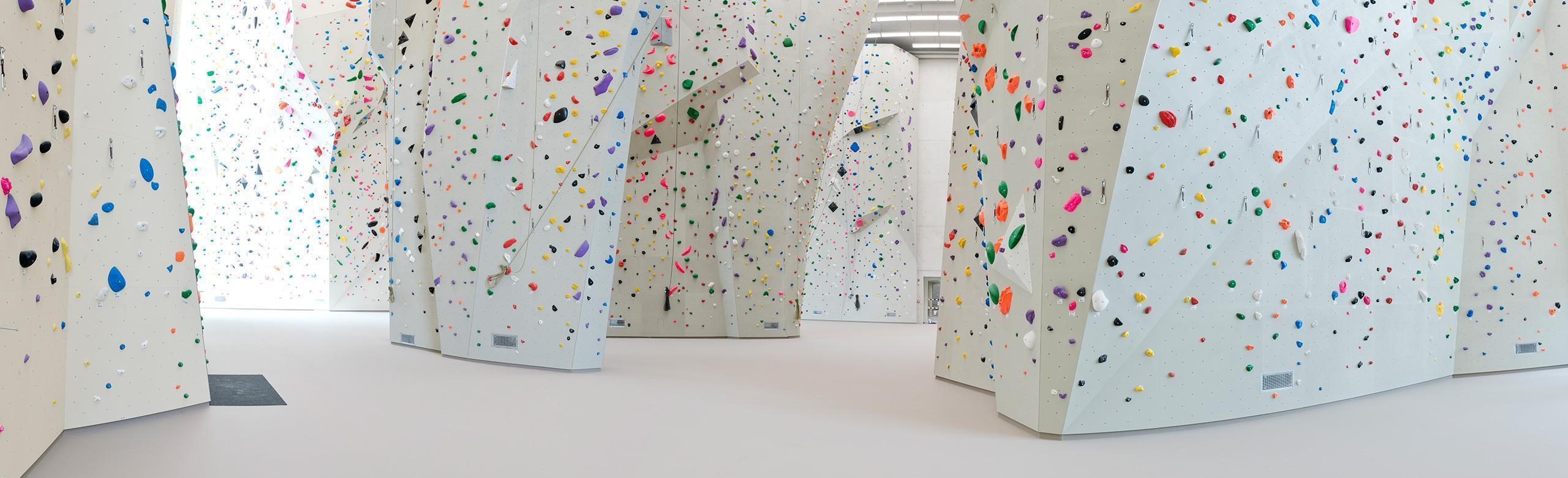 A light-coloured floor covering in a climbing hall plus various white climbing walls with different coloured holds and routes. 