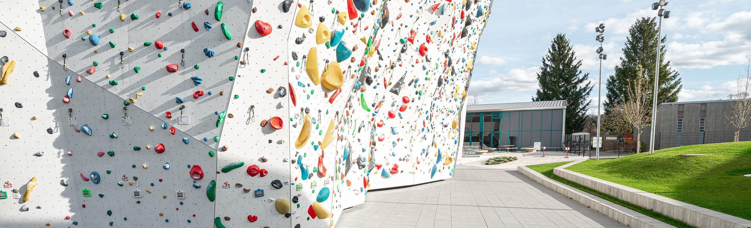 Outdoor climbing wall with fall protection floor A light-coloured floor covering in the outdoor area of a climbing hall. The flooring lies directly under the climbing wall. In the background are trees and a green meadow.