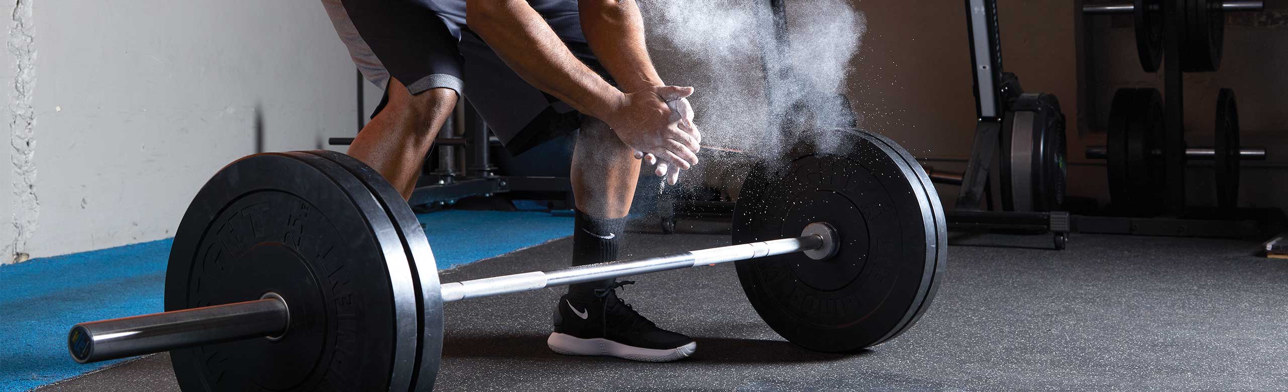 A man rubbing his hands with chalk. In front of him is a barbell lying on the REGUPOL floor covering. The grey flooring provides acoustic benefits in a fitness studio, even with falling barbells.