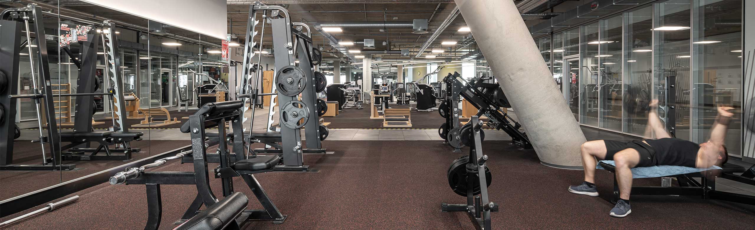 REGUPOL flooring in an area for free weight training in a fitness centre. You can see a man lying on a weight bench lifting weights.