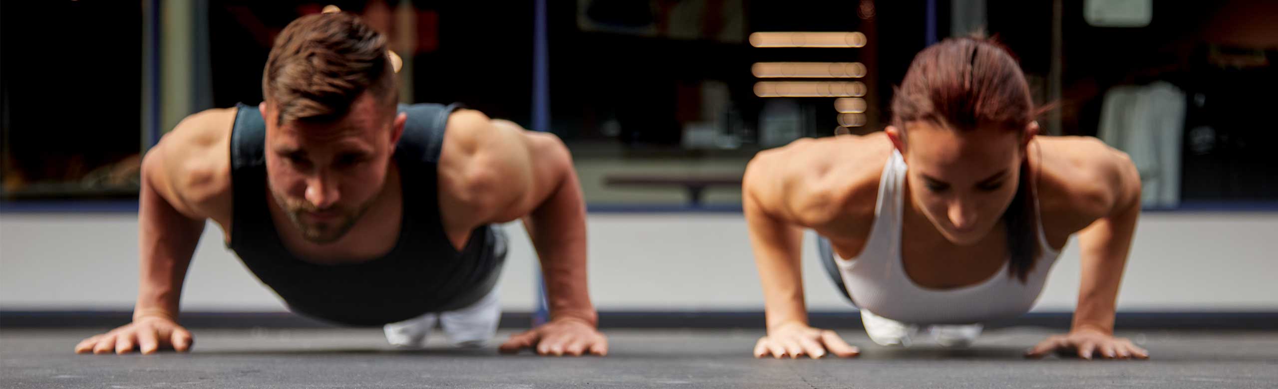 A man and a woman do push-ups on a REGUPOL flooring. A man and a woman doing push-ups on a REGUPOL floor covering. The floor is grey. Mirrors in the fitness room can be seen in the background.