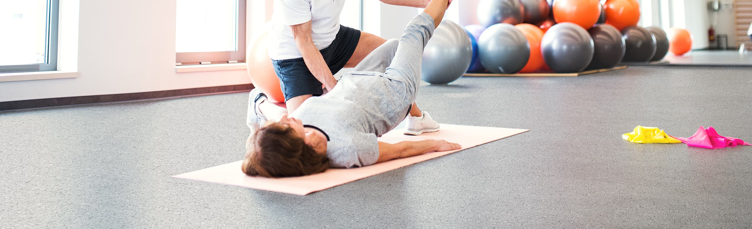 Targeted physio training support in the gym with a trainer A woman lies on a REGUPOL mat in the fitness room and lifts one leg while a trainer supports her during a physio exercise. Exercise balls and fitness bands can be seen in the background.