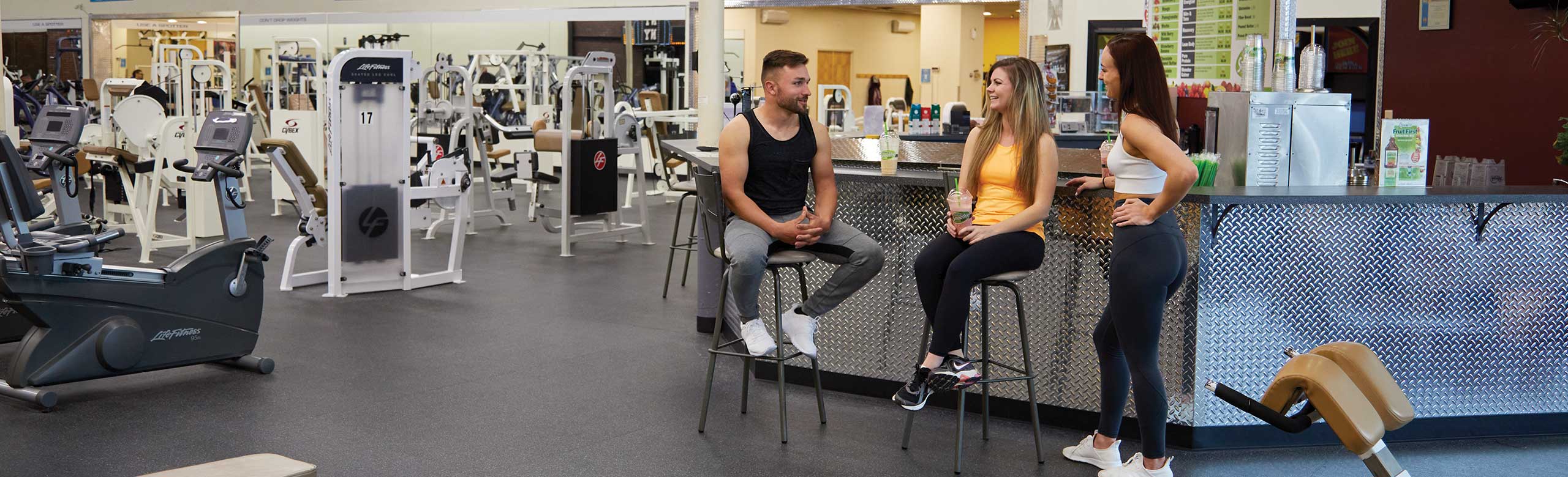 Two women and a man are chatting at a bar in the gym.  Fitness equipment can be seen in the background. The REGUPOL floor covering has been chosen in dark grey.