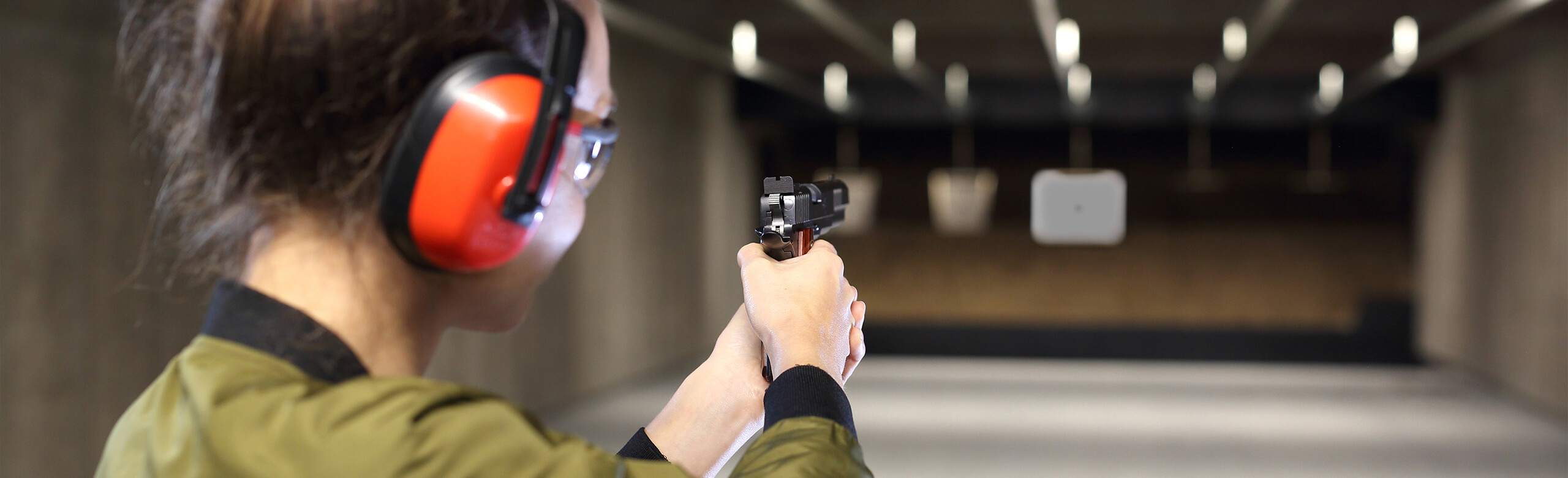 Woman with protective goggles and protective earphones aims a gun at a target in a bullet trap in a shooting range