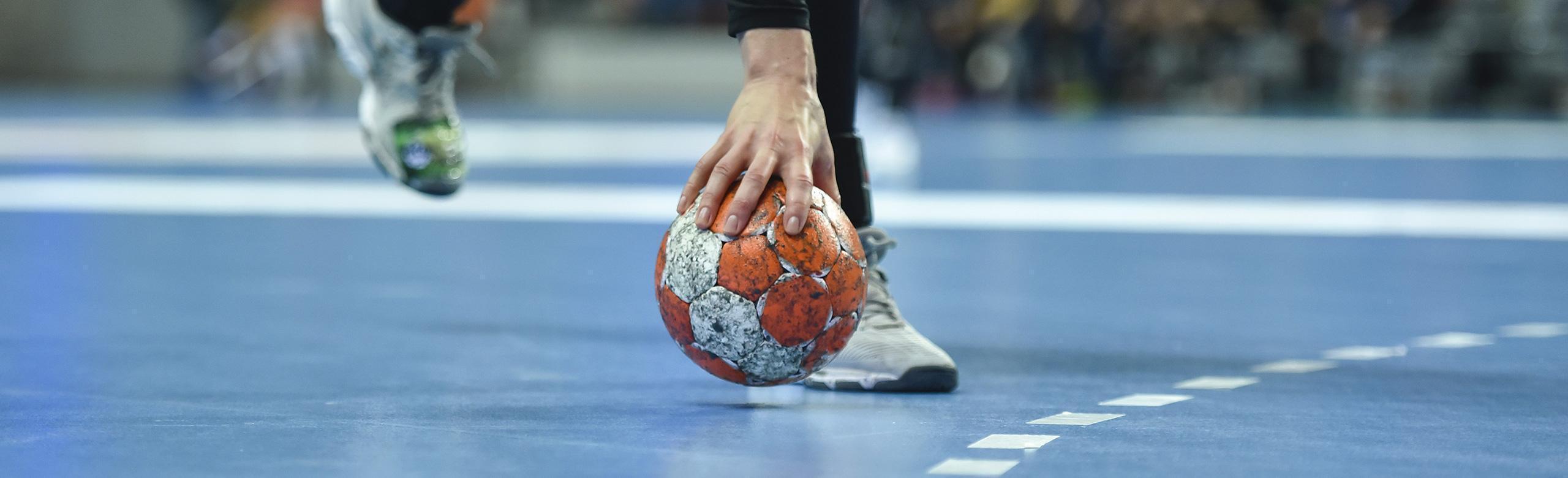 An athlete reaches for a handball in a sports hall.  The flooring of the sports hall is blue, white lines mark the playing field. 