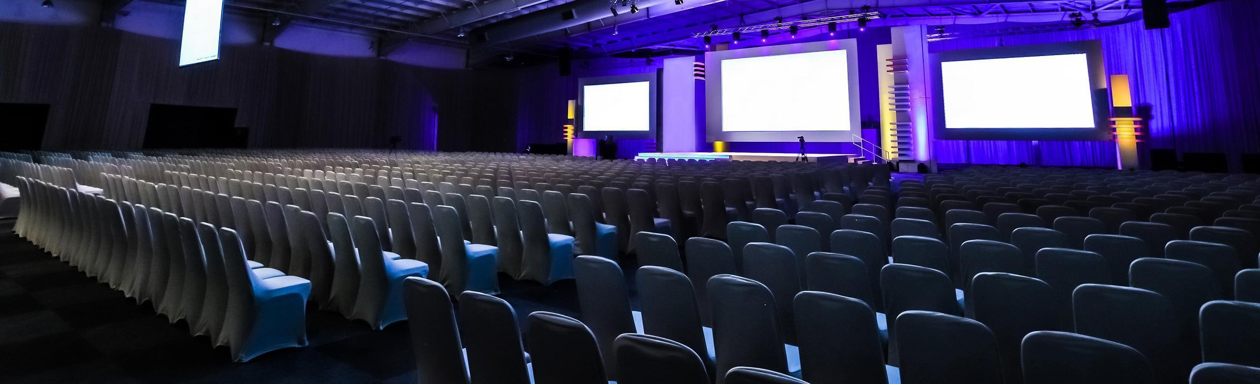 Many rows of chairs pointing towards the stage.  The stage is located in a sports hall and is backlit with purple-coloured light. Three large screens hang on the stage wall. 