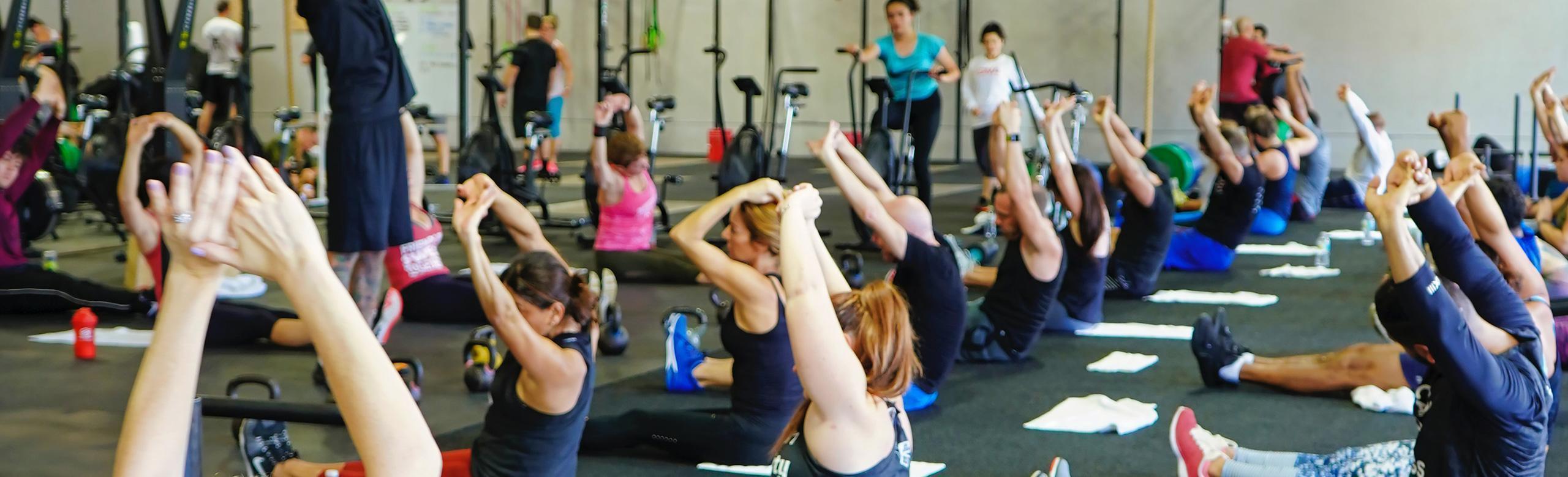 Several people are doing group sports in a room.  They sit on the floor and stretch their arms upwards. Some have towels and drinking bottles on the floor. In the background you can see some fitness equipment for group training. 