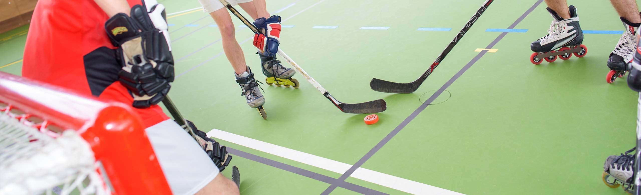 Roller sports in a sports hall Various people are playing inline hockey in a sports hall on a green sports hall floor with different lines. They are wearing inline skates and hockey gloves, have hockey sticks in their hands and pass the puck to each other in front of a goal.