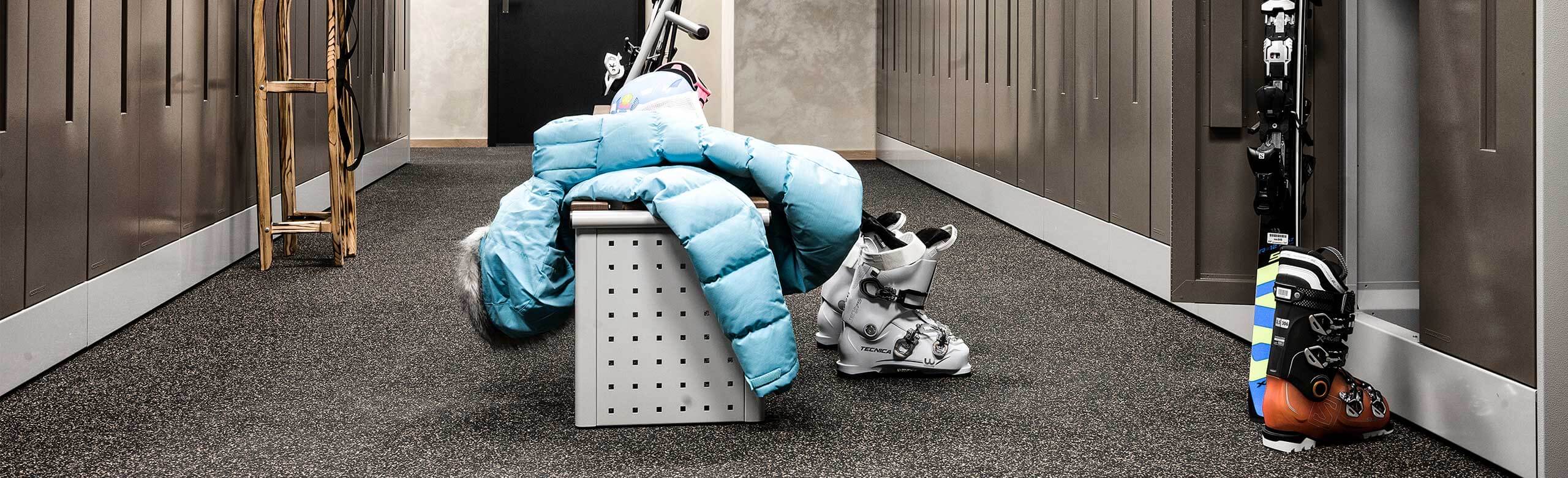 A room for ski equipment in a hotel. You can see ski boots and skis, various lockers and a seat with a jacket on it. The floor covering is dark grey.