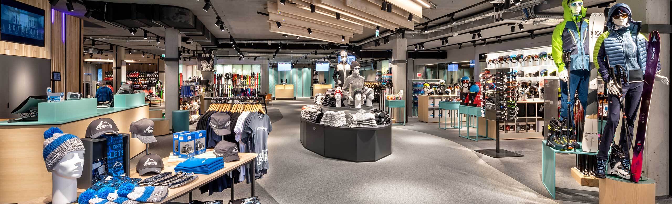A modern ski shop in a mountain railway station. Lots of tables and dolls with ski equipment and ski accessories. The flooring is two-coloured, one area is light grey and the other is dark grey.