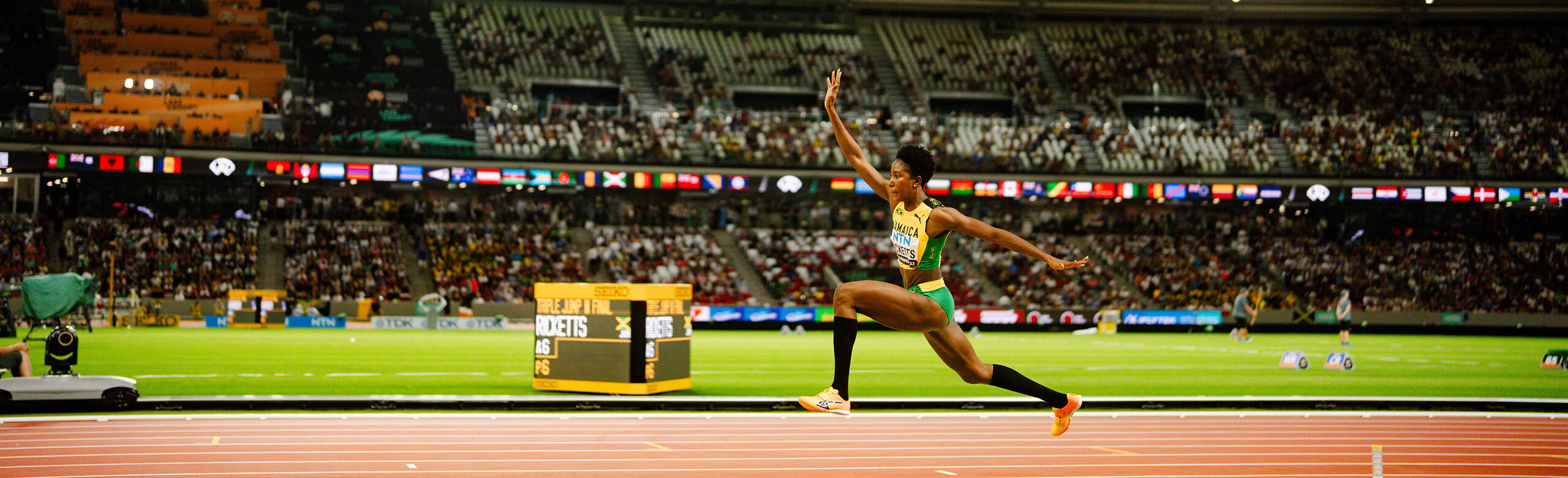 Triple jump by a female athlete at an international floodlit athletics event on a synthetic runway. Female athlete mid-flight during the hop phase of a triple jump at an international athletics competition in a packed stadium. The runway features color-marked synthetic surfacing under stadium floodlights.