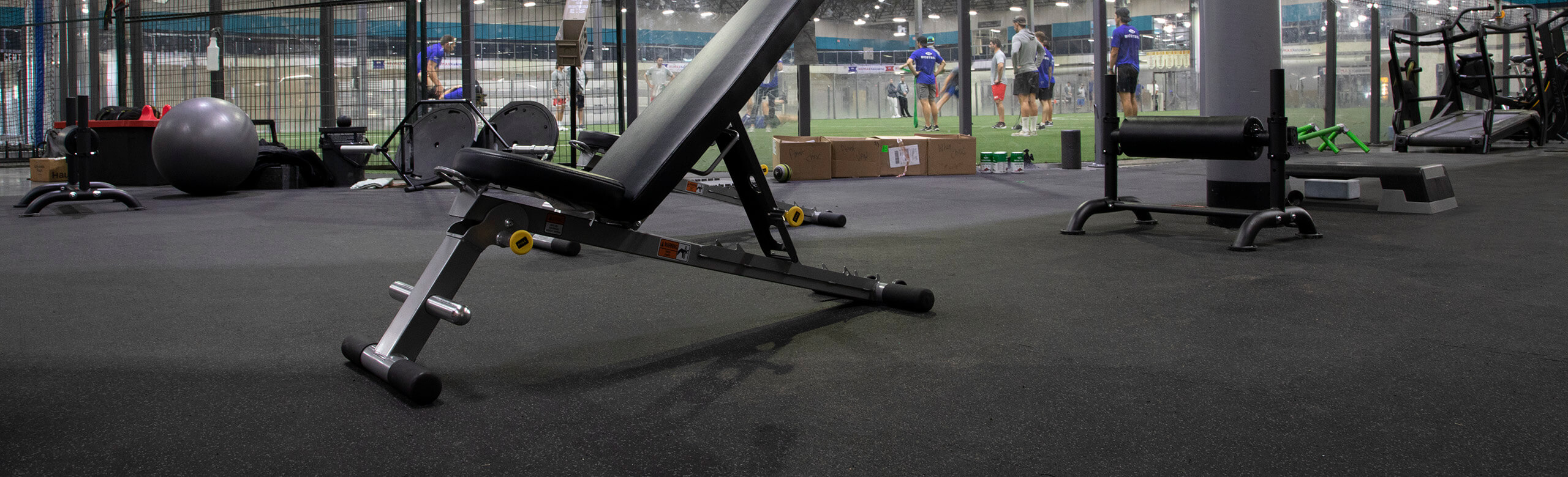 Kelowna High Performance - Weight Bench & REGUPOL aktiv Flooring A inclined weight lifting bench rests on REGUPO aktiv rolled rubber flooring, with a view of the turf area in the background of Kelowna High Performance. There are a number of athletes located on the turf, receiving instruction from a coach.