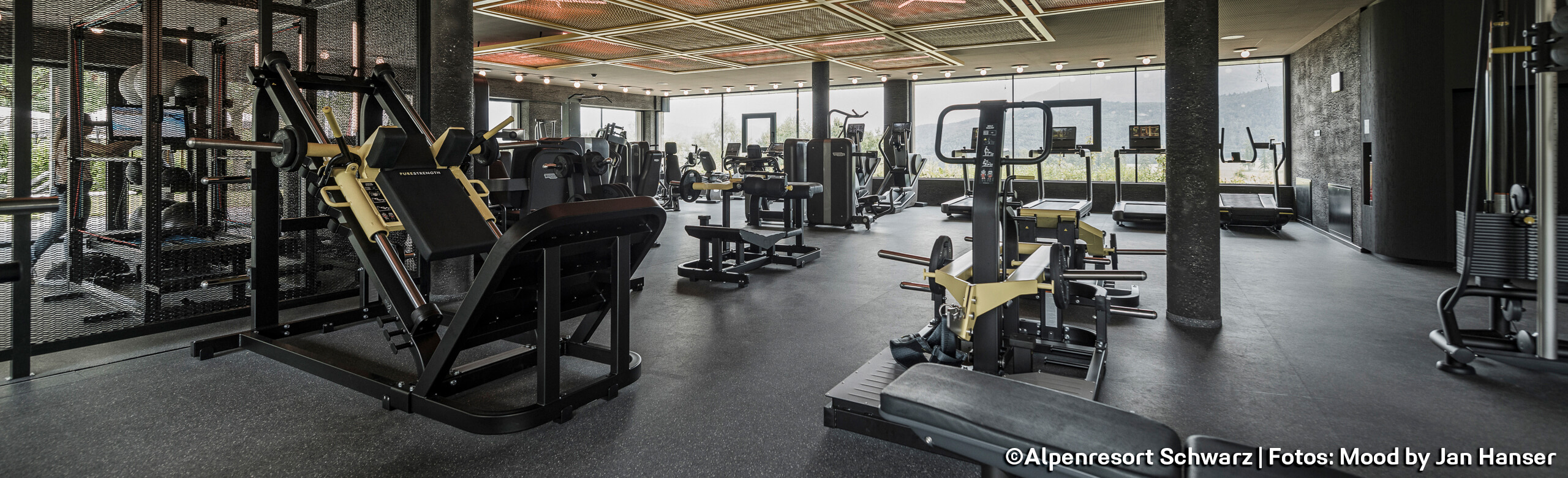 A fitness studio in a hotel with lots of training equipment and a dark-coloured floor. There is a large window at the end of the room.